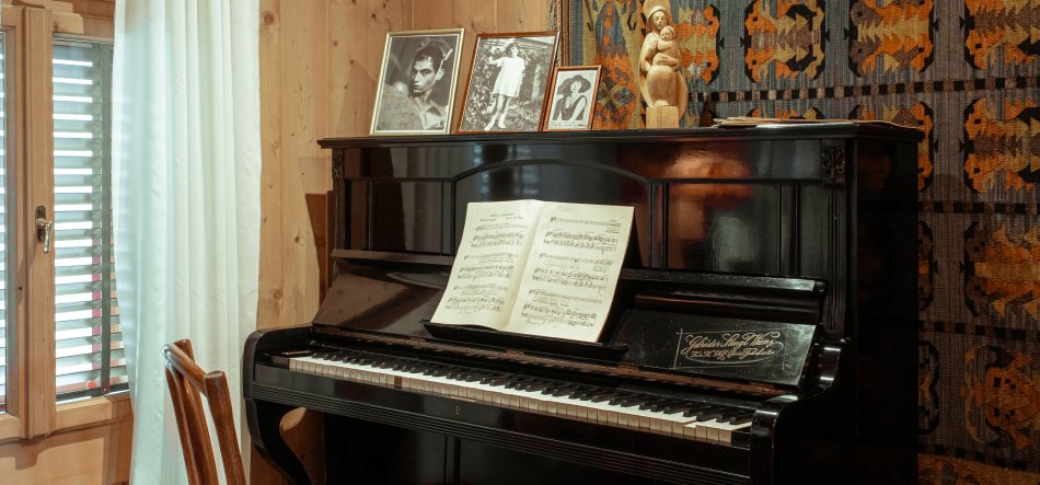 Czarne, lakierowane pianino przy drewnianej ścianie z otwartą nutą na pulpicie, trzema oprawionymi zdjęciami i drewnianą rzeźbą na wieczku, obok okno z białą zasłoną i drewniane krzesło, a za instrumentem wiszący kolorowy, geometryczny gobelin.