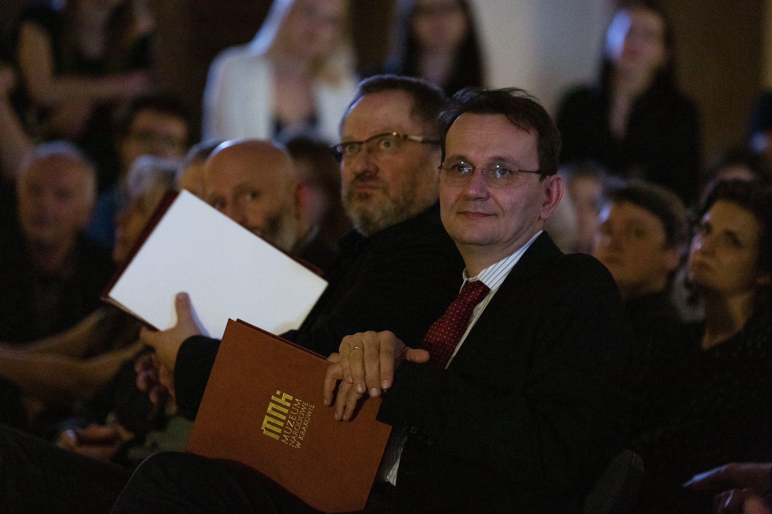 Mężczyzna w okularach w garniturze i czerwonym krawacie siedzi na widowni i trzyma brązową teczkę z złotym napisem, za nim rozmyte postacie innych uczestników w przyciemnionym pomieszczeniu.