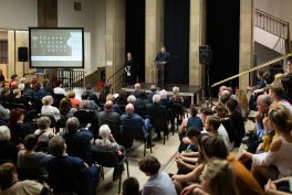 Wnętrze dużej sali z publicznością siedzącą i stojącą przed niewielką sceną, na której stoją dwie osoby przy podium; po lewej ekran projekcyjny, widoczne kolumny, głośniki i schody po prawej.