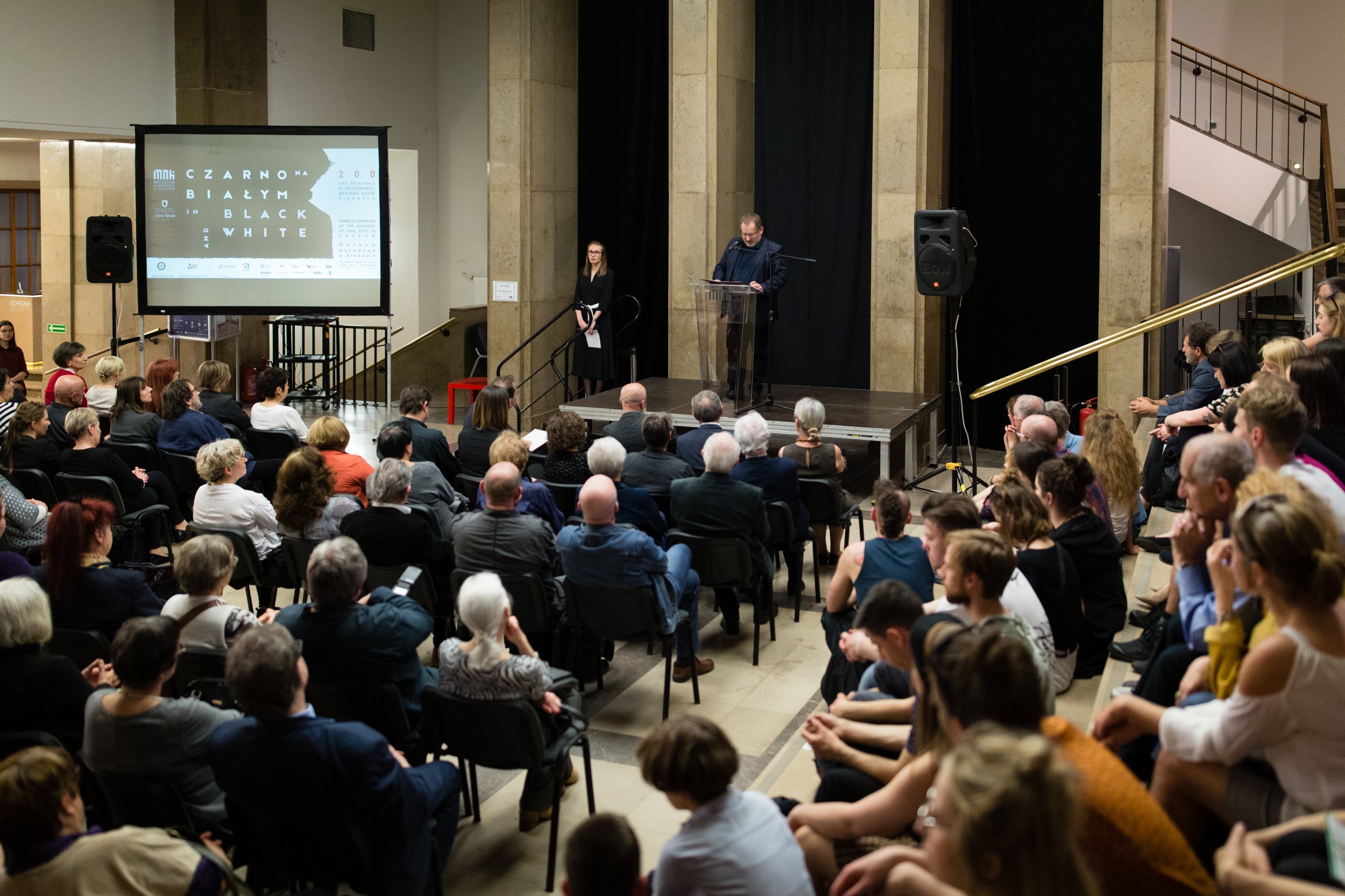Duża sala z publicznością siedzącą w rzędach i na schodach, mężczyzna przemawiający przy przezroczystym pulpicie na niewielkiej scenie przed czarnym tłem, stojąca obok kobieta, po lewej ekran projekcyjny z napisem „CZARNO BIAŁYM / IN BLACK AND WHITE” oraz widoczne kamienne kolumny i głośniki.