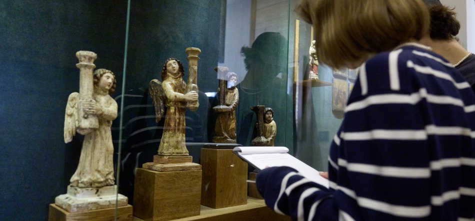 Osoba w granatowo‑białej bluzce w paski trzyma notatnik i ogląda przez szkło gabloty kilka drewnianych, polichromowanych rzeźb aniołów ustawionych na drewnianych postumentach na zielonym tle; obok widać częściowo drugą osobę.