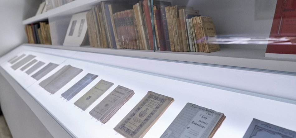 Podłużna, podświetlona gablotka wystawowa z ułożonymi w rzędzie pożółkłymi książeczkami i broszurami na płaskiej powierzchni oraz półką za szkłem z ustawionymi tomami w tle, sfotografowana pod kątem.