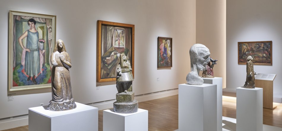 Wnętrze jasnej galerii z białymi ścianami i drewnianą podłogą, na białych piedestałach ustawione kilka rzeźb — po lewej stylizowana figura kobiety, obok geometryczna wielopoziomowa forma, dalej duża rzeźba głowy i mniejsza stojąca postać — a na ścianach wiszą kolorowe obrazy w ramach.