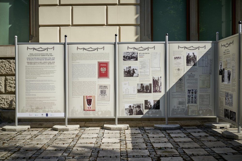 Kilka stojących białych paneli informacyjnych z tekstem, czarno‑białymi fotografiami i kolorowymi plakatami osadzonych w metalowych ramach na betonowych podstawach przed elewacją budynku, ustawionych na kamiennym placu, z ukośnym cieniem padającym na część paneli.