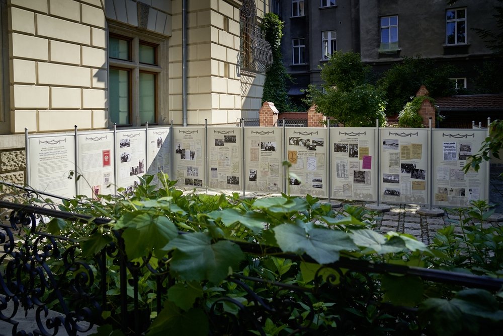 Dziedziniec z rzędem białych tablic informacyjnych z dokumentami i czarno-białymi zdjęciami ustawionych przed jasną elewacją kamienicy, w pierwszym planie ozdobna żelazna balustrada i bujne zielone liście, a w tle ciemniejsza fasada sąsiedniego budynku i drzewa.