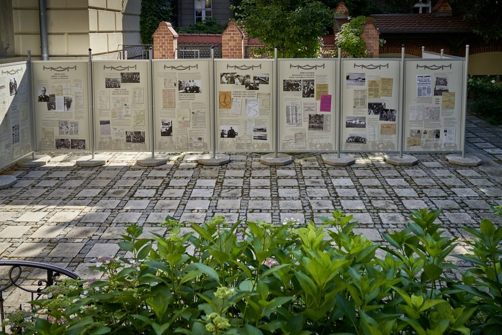Rząd stojących paneli informacyjnych z dokumentami i czarno‑białymi zdjęciami ustawionych na betonowych podstawach na brukowanym dziedzińcu, z zielonymi krzewami na pierwszym planie i ceglaną zabudową oraz drzewami w tle.
