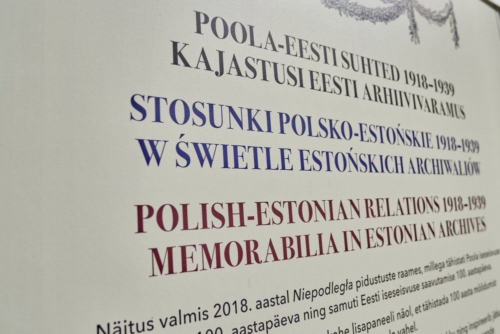 Zbliżenie na biały panel z poziomymi tytułami w trzech językach: czarny estoński napis u góry, duży niebieski polski napis STOSUNKI POLSKO-ESTOŃSKIE 1918–1939 pośrodku, bordowy angielski napis POLISH-ESTONIAN RELATIONS 1918–1939 MEMORABILIA IN ESTONIAN ARCHIVES poniżej oraz fragment drobnego tekstu informacyjnego na dole i dekoracyjny wieniec w prawym górnym rogu.