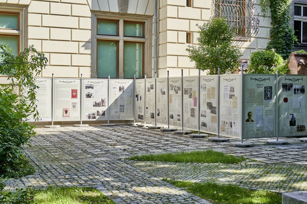 Dziedziniec z brukowaną nawierzchnią i trawnikami, na którym ustawiono rząd wolnostojących paneli wystawowych z tekstami i zdjęciami przed jasną elewacją budynku z dużymi oknami, wokół krzewy, drzewa i cieniste plamy światła.