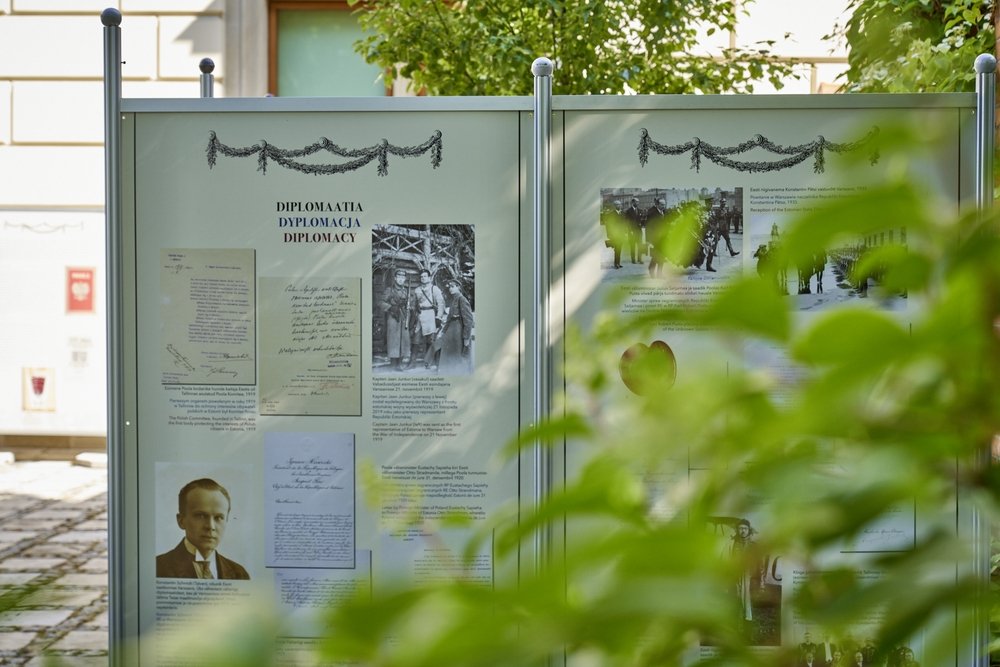 Przed budynkiem ustawione dwie metalowe, przeszklone tablice wystawowe z dokumentami, czarno-białymi zdjęciami i napisem "DIPLOMAATIA DYPLOMACJA DIPLOMACY" pod dekoracyjnymi girlandami, częściowo zasłonięte zielonymi liśćmi na pierwszym planie.