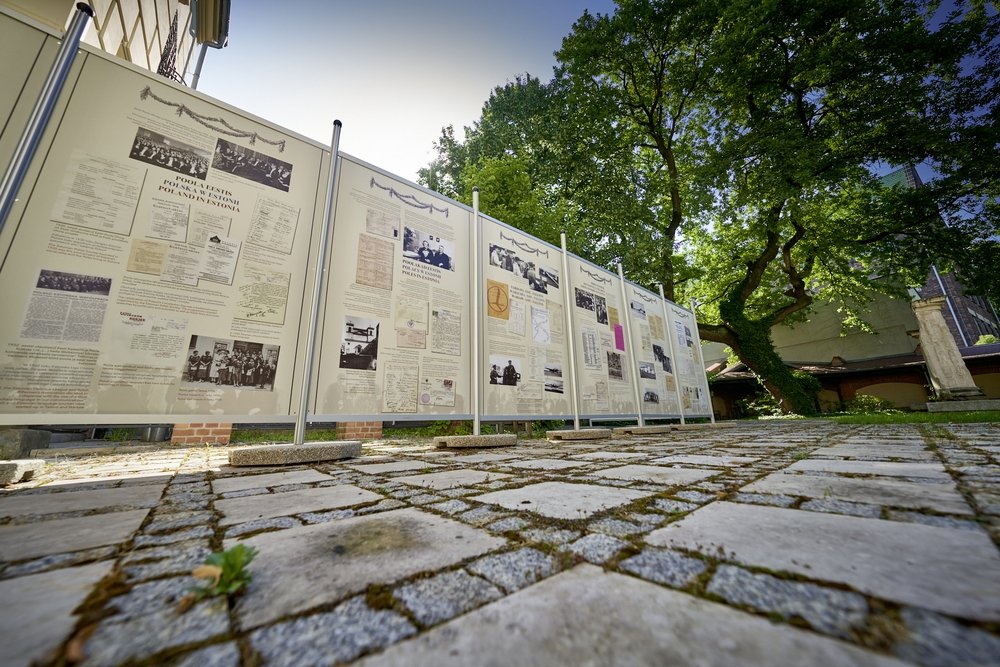 Niski kąt widzenia na zewnętrzną wystawę kilkunastu tablic informacyjnych z reprodukcjami dokumentów i zdjęć zamocowanych na metalowych ramach ustawionych w rzędzie na brukowanym, porośniętym mchem podwórzu, w tle duże drzewa i fragmenty budynków.