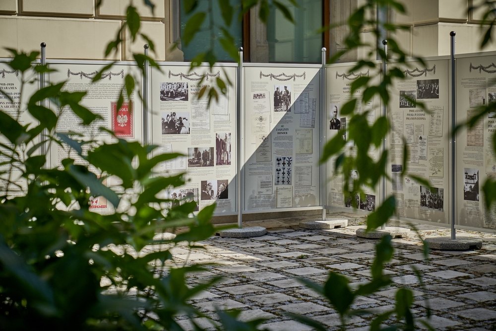 Widok przez rozmyte zielone liście na zewnętrzną wystawę tablic informacyjnych z zdjęciami i dokumentami, zamocowanych na metalowych słupkach z betonowymi podstawami na brukowanej nawierzchni przed ścianą budynku z oknem.
