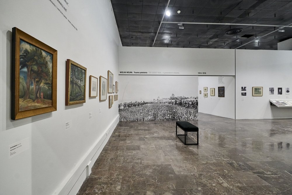 Wnętrze galerii z białymi ścianami, po lewej linia oprawionych obrazów i grafik, na tylnej ścianie duża czarno-biała fotografia pola z żołnierzami i napisem WIELKA WOJNA 1914–1918, na środku czarna ławka i błyszcząca kamienna posadzka.