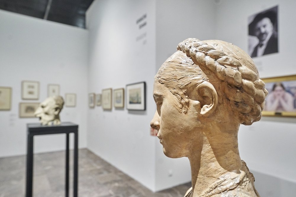 Prawy profil ceramicznego popiersia kobiety z warkoczem na pierwszym planie w galerii, w tle rozmyta rzeźba na czarnym postumencie oraz oprawione rysunki i fotografie na białych ścianach.