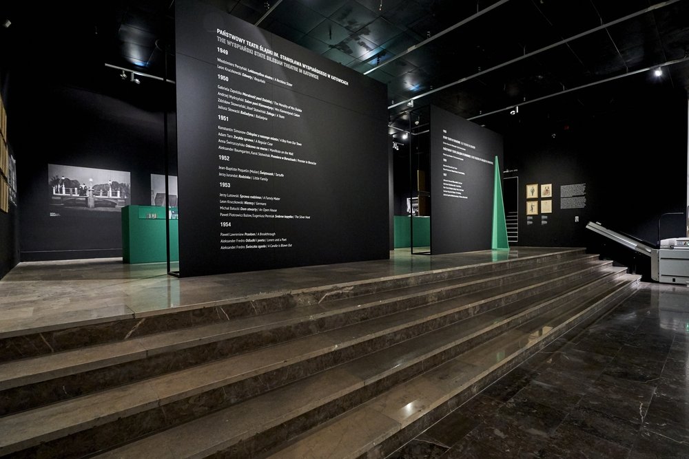 Ciemne wnętrze galerii z szerokimi marmurowymi schodami na pierwszym planie, czarnymi ścianami z dużymi białymi napisami informacyjnymi, zielonymi gablotami i oprawionymi fotografiami przy punktowym oświetleniu.