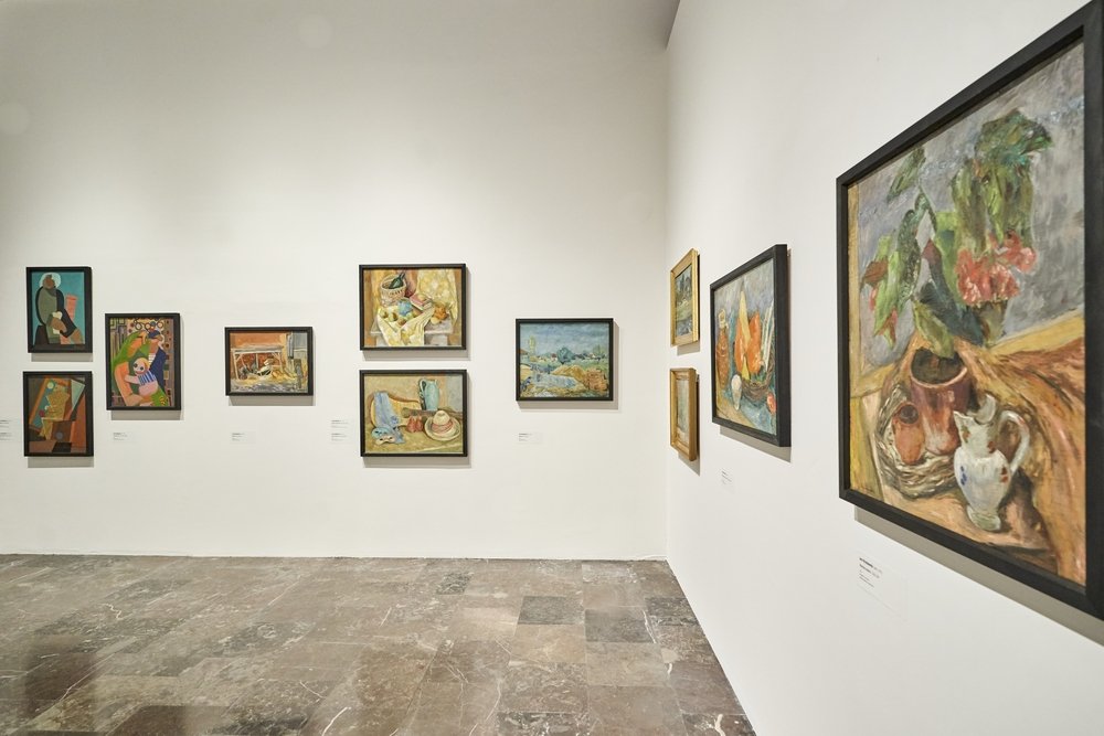 Widok wnętrza galerii z białymi ścianami i marmurową podłogą, na których w rzędach powieszono oprawione obrazy — po lewej i na wprost kilka małych i średnich prac przedstawiających portrety, pejzaże i martwe natury, a po prawej duży obraz z bukietem kwiatów i naczyniami.