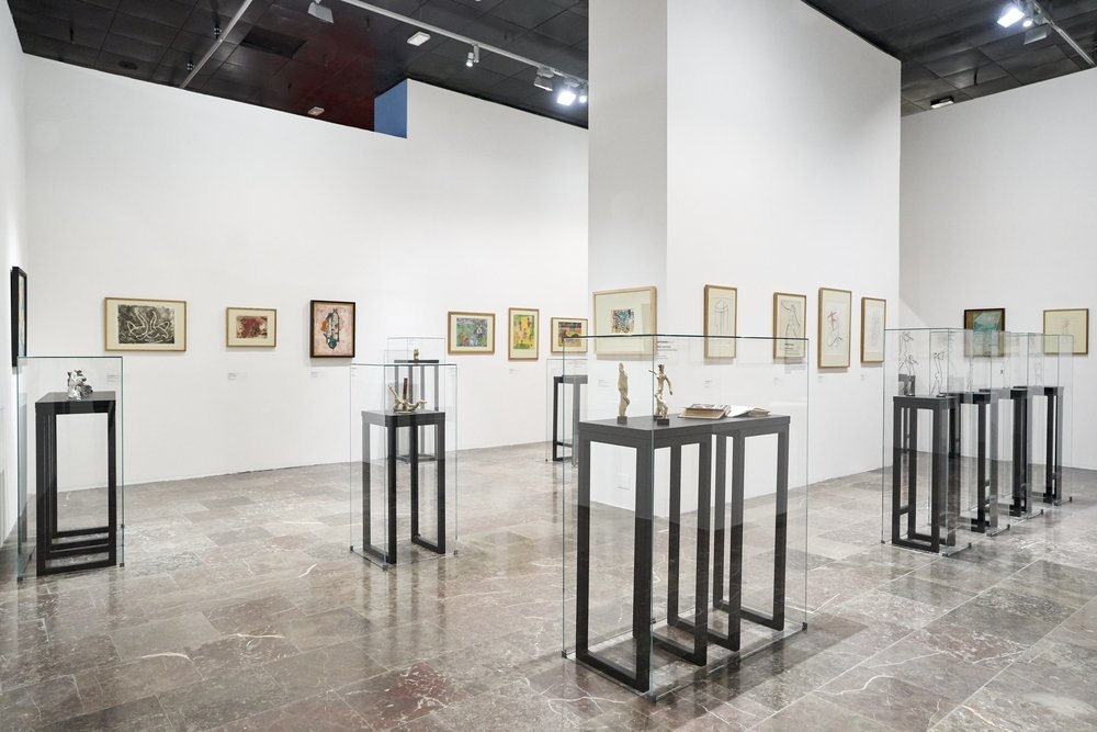 Wnętrze jasnej galerii sztuki z białymi ścianami i wysokim sufitem, na których wiszą oprawione rysunki i obrazy, oraz kilkoma przeszklonymi gablotami na czarnych metalowych postumentach wystawiającymi małe rzeźby, z centralnym białym filarem i marmurową posadzką.