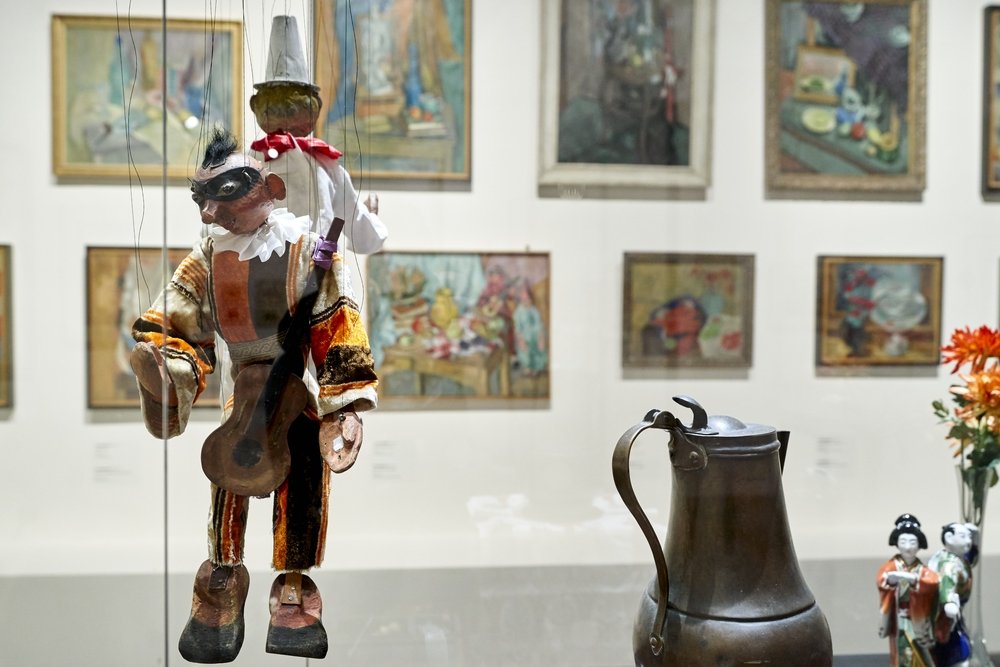 Marionetka na sznurkach w kolorowym, pasiastym kostiumie i czarnej masce trzymająca małą gitarę na pierwszym planie, obok metalowy dzbanek, drobne figurki i bukiet kwiatów, a w tle ściana z wieloma oprawionymi obrazami.
