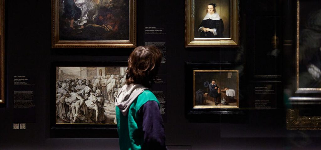Osoba widziana od tyłu w zielono‑granatowej bluzie z kapturem stoi przed ciemną ścianą wystawową z kilkoma oprawionymi w złote ramy obrazami — dużą sceną grupową, portretem kobiety i mniejszym przedstawieniem mężczyzny przy stole — oraz widocznymi tabliczkami informacyjnymi.