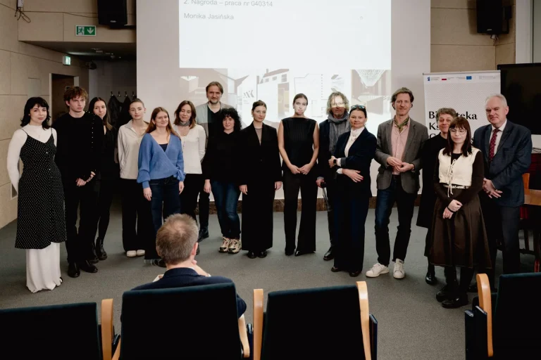 Grupowe zdjęcie kilkunastu osób ustawionych w jednym rzędzie przed dużym białym ekranem projekcyjnym w sali konferencyjnej, na pierwszym planie fotele i tył głowy siedzącego widza, po prawej baner z napisem "Biblioteka", po lewej drzwi i zielony znak wyjścia ewakuacyjnego.