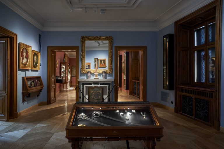 Galeria muzealna z niebieskimi ścianami i drewnianymi framugami: na pierwszym planie oszklona gablotka z mieczami i medalami, w centrum złota ornamentowana rama lustra nad marmurowym kominkiem z dwiema zdobionymi wazonami i rośniętym panelem przed kominkiem, po bokach drzwi prowadzące do sąsiednich sal z różowymi ścianami i meblami ekspozycyjnymi oraz liczne portrety w złotych ramach, z prawej okno z ozdobnymi szybami.
