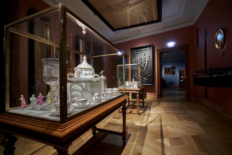 Sala wystawowa z przeszklonymi gablotami na drewnianych stolikach prezentującymi ozdobną porcelanową zastawę, drobne figurki i kryształowe naczynie, czerwone ściany, szklana gablotka ze zbiorami szabli na ścianie oraz przejście do sąsiedniego, niebiesko oświetlonego pokoju z obrazami.