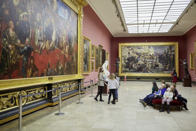 Wnętrze sali galerii z wysokim świetlikiem, czerwonymi ścianami i wielkimi złoconymi ramami z obrazami przedstawiającymi liczne postaci, ozdobną czarno‑złotą podstawą pod jednym z obrazów, marmurową rzeźbą i ciemną statuą przy ścianie, barierkami chroniącymi eksponaty oraz grupą dzieci siedzących na okrągłej czerwonej ławce i kilkoma osobami stojącymi obok.
