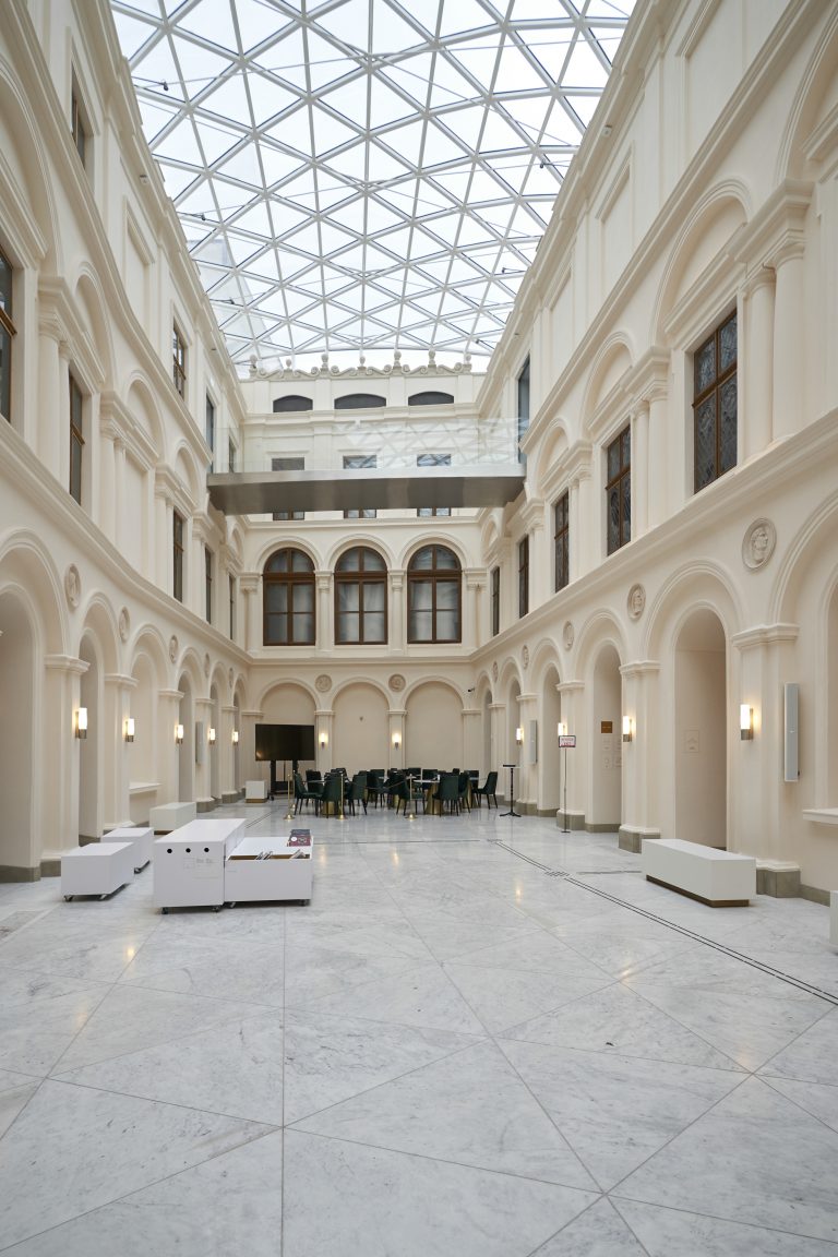 Wnętrze jasnego, neoklasycznego atrium z wysokimi kremowymi ścianami ozdobionymi łukami i pilastrami, rzędami drewnianych okien, geometrycznym szklanym dachem, zawieszoną metalową kładką, marmurową posadzką oraz stolikami z zielonymi krzesłami w centrum i białymi siedziskami oraz kinkietami wzdłuż boków.