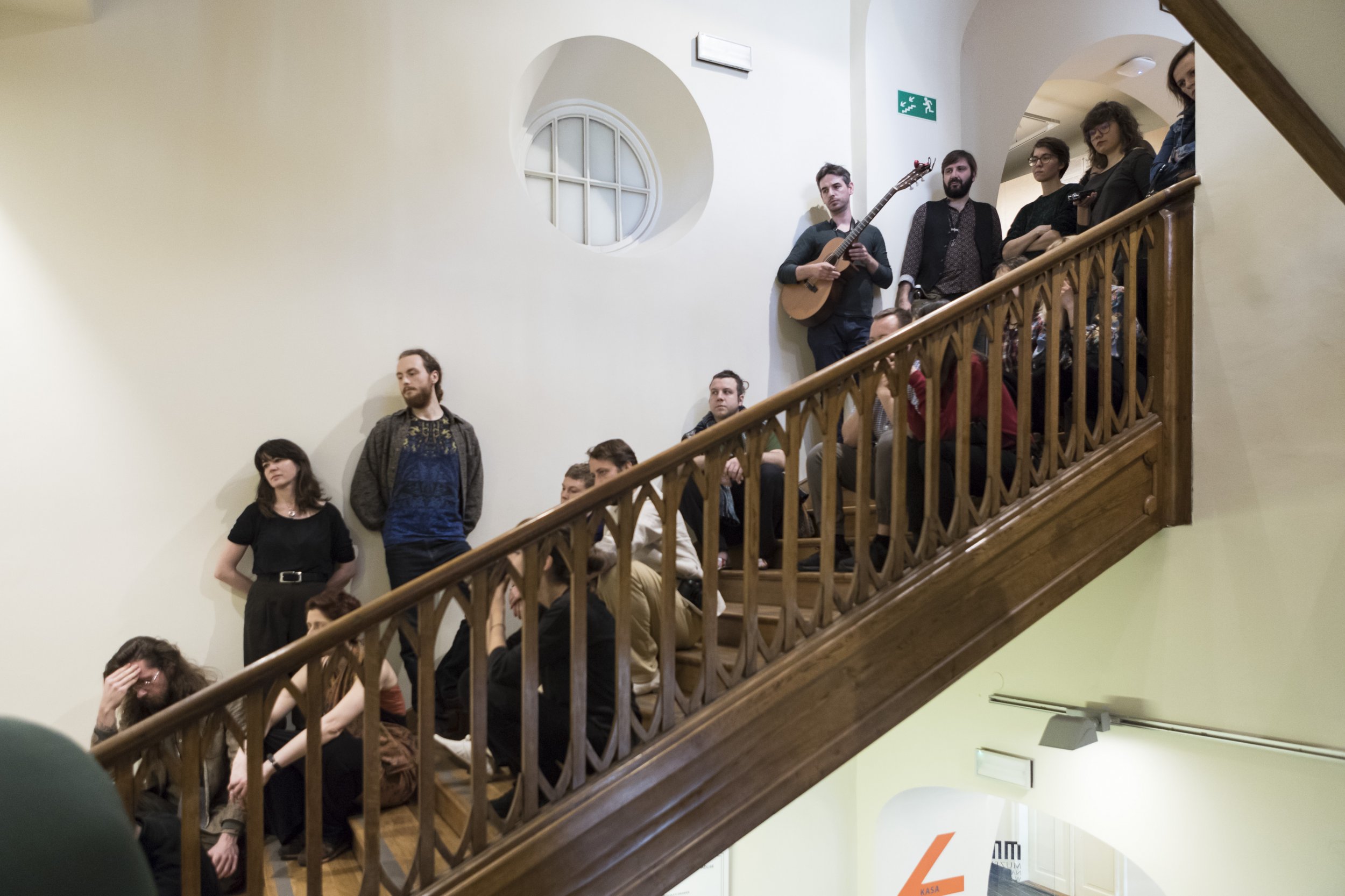 Grupa osób siedzących i stojących na drewnianych schodach z ozdobną balustradą wewnątrz jasnego budynku; u góry po prawej osoba z gitarą przy okrągłym oknie, na ścianie zielony znak ewakuacyjny i przez przejście widoczny fragment szyldu z pomarańczowym elementem.