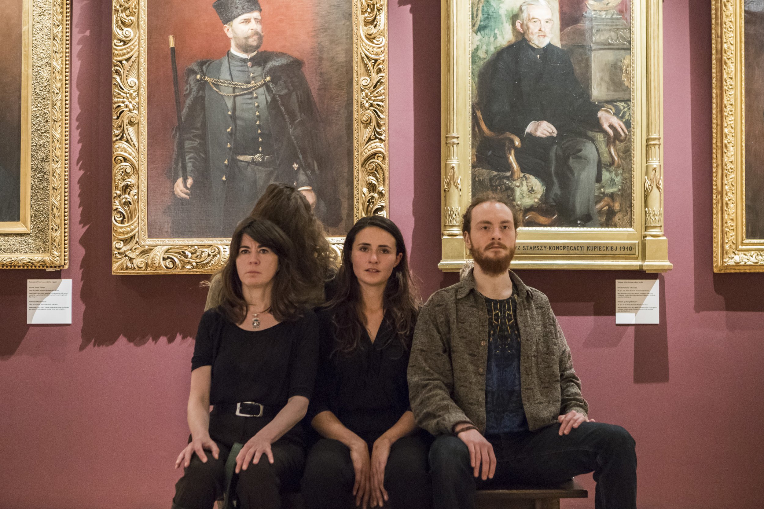 Dwie kobiety i mężczyzna siedzą obok siebie na ławce przed ciemnoróżową ścianą galerii sztuki, a za nimi wiszą duże olejne portrety mężczyzn w ozdobnych pozłacanych ramach z widocznymi tabliczkami informacyjnymi.
