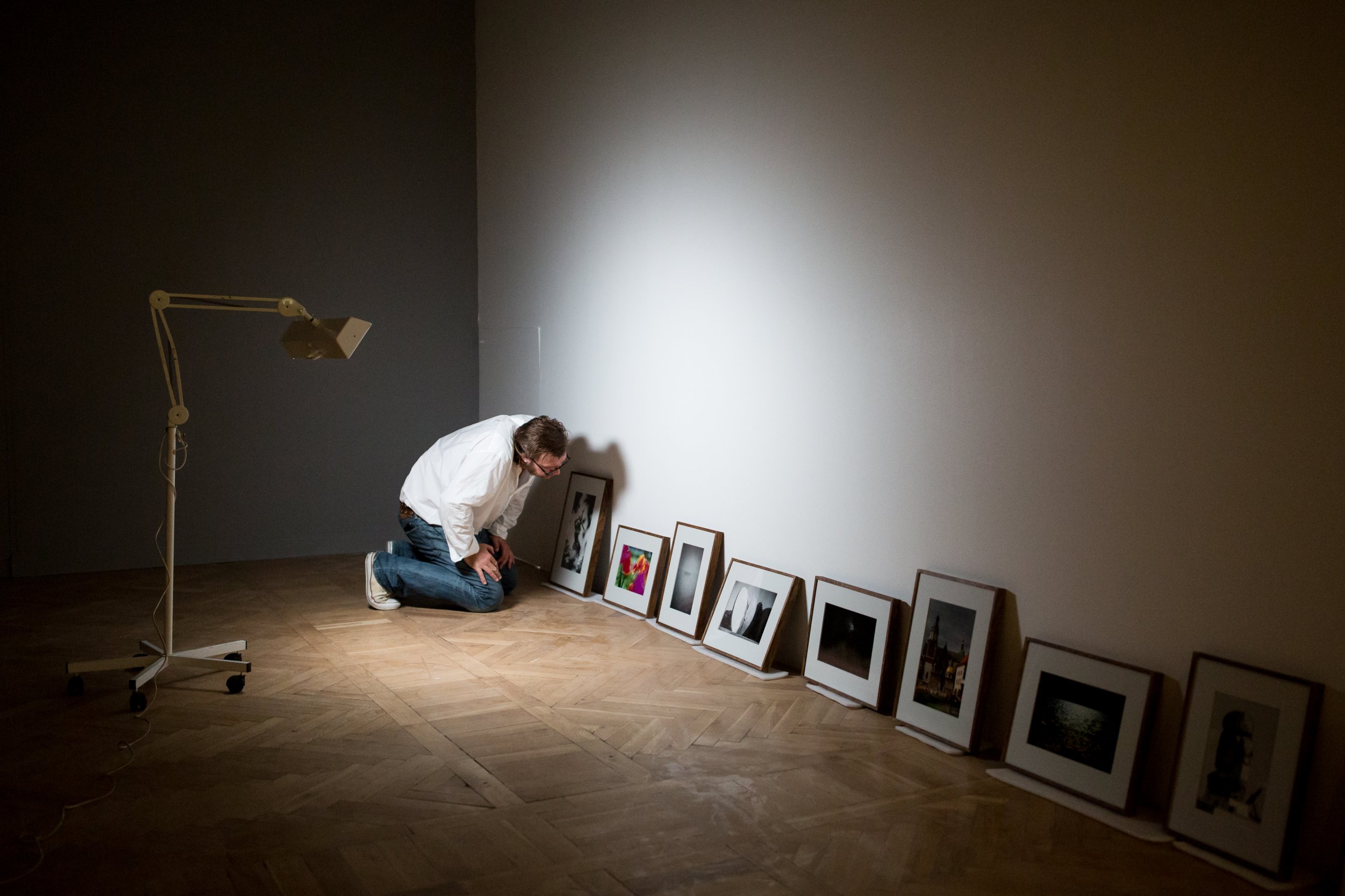 W ciemnym pomieszczeniu z drewnianą podłogą lampa na ruchomej nodze po lewej oświetla osobę w białej koszuli i dżinsach, klęczącą i pochyloną nad szeregiem oprawionych zdjęć i obrazów ustawionych pod ścianą.