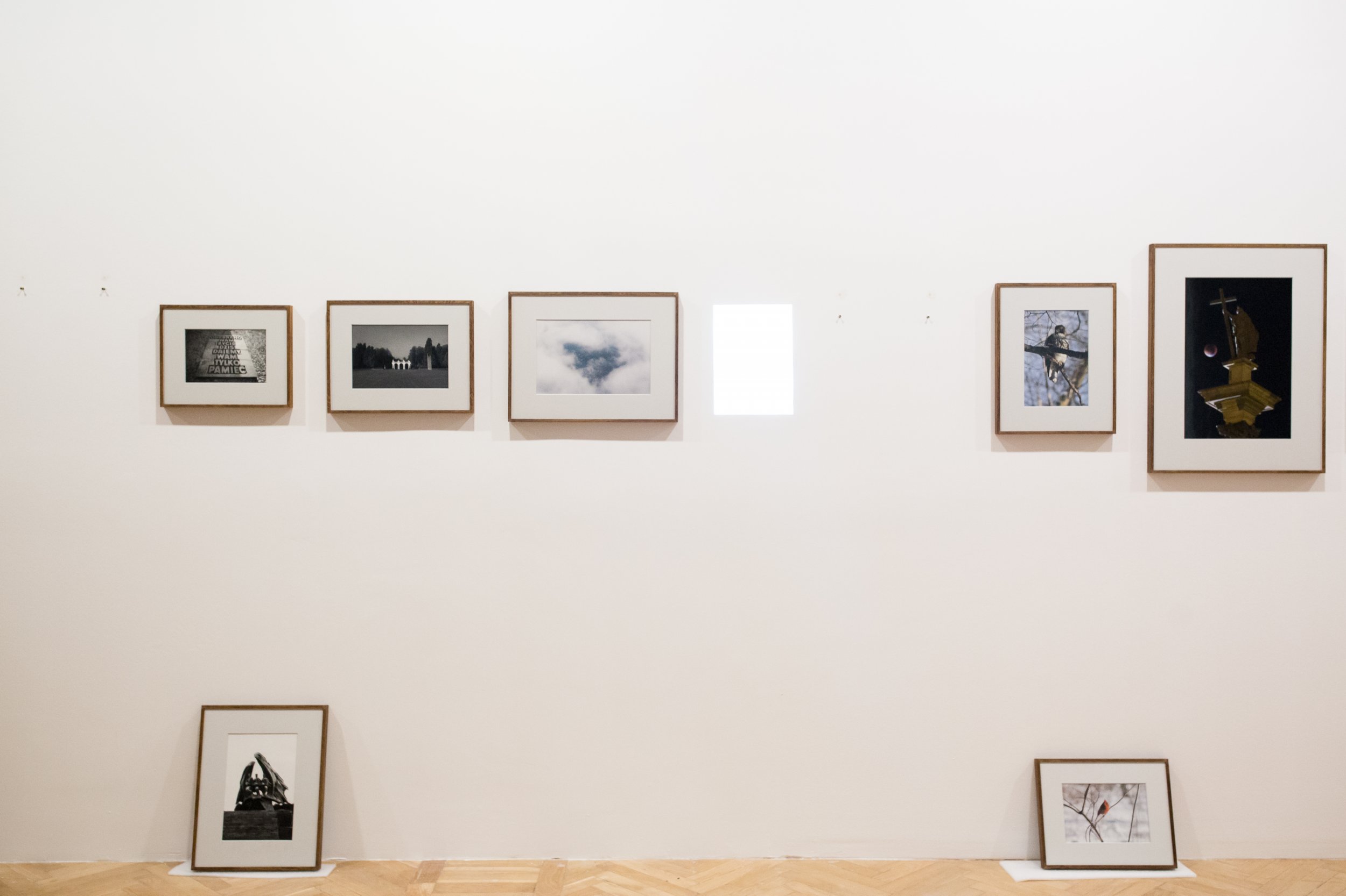 Biała ściana galerii z szeregiem oprawionych fotografii i grafik w cienkich drewnianych ramach zawieszonych wysoko (pięć ramek od lewej do prawej z jasnym prostokątem światła między trzecią a czwartą) oraz dwiema ramami opartymi o jodełkową drewnianą podłogę po lewej i prawej.
