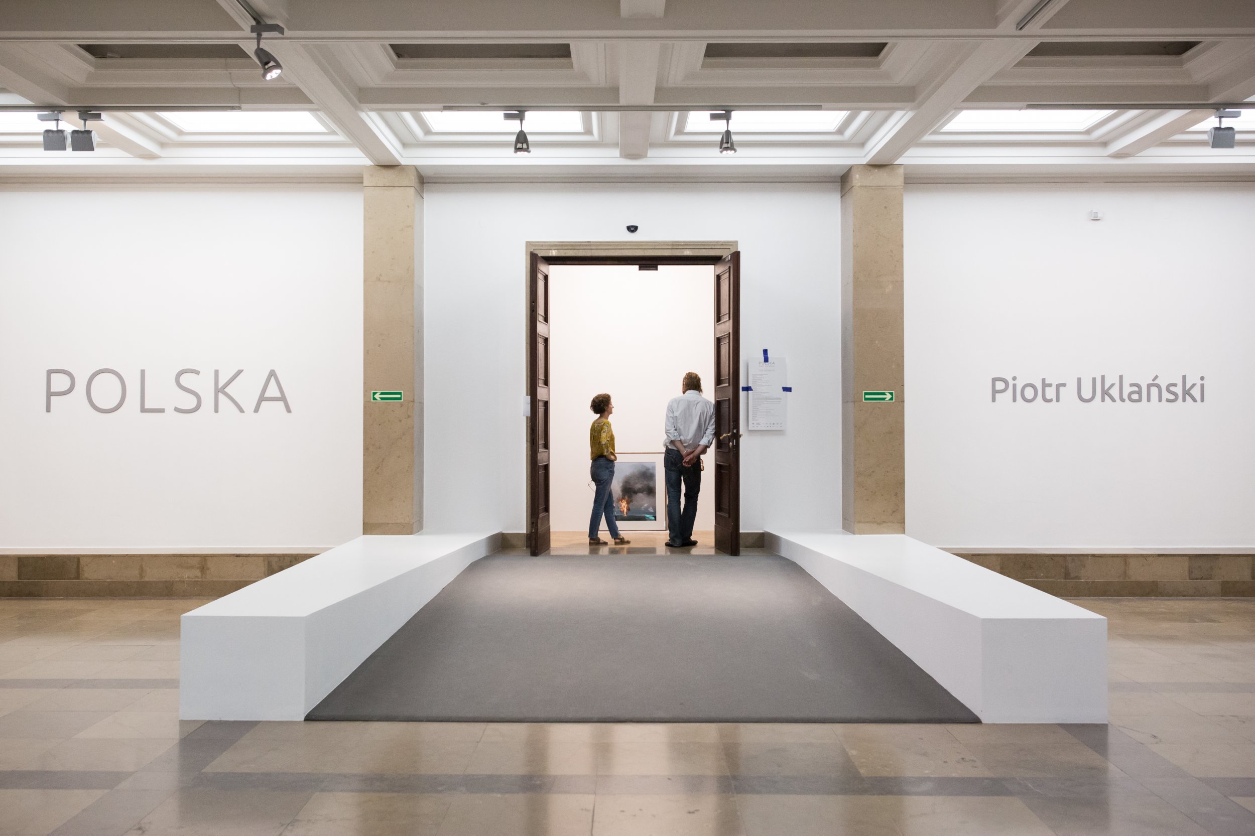 Wnętrze galerii z centralnym otwartym wejściem i drewnianymi drzwiami, po bokach białe ściany z napisem "POLSKA" po lewej i "Piotr Uklański" po prawej, dwie osoby stojące w drzwiach oglądające obraz we wnętrzu, niskie białe ławy lub rampy po obu stronach, marmurowe filary, zielone znaki ewakuacyjne oraz sufit ze świetlikami i reflektorami.