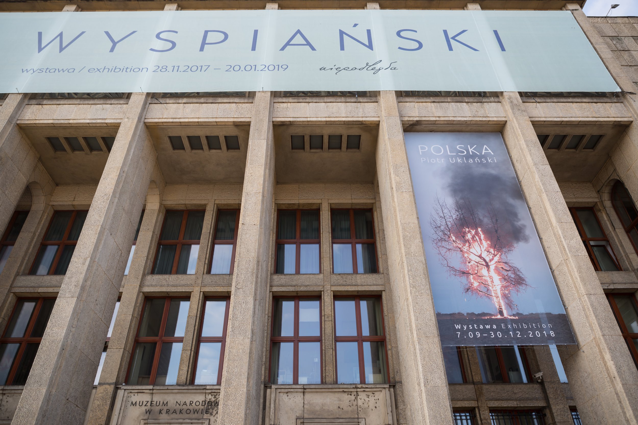 Fasada budynku z wysokimi betonowymi kolumnami i wieloma prostokątnymi oknami, u góry duży poziomy baner z napisem „WYSPIAŃSKI” i datami wystawy, po prawej pionowy baner z fotografią płonącego drzewa i napisem „POLSKA Piotr Uklański”, a niżej częściowo widoczny kamienny napis „Muzeum Narodowe w Krakowie”.