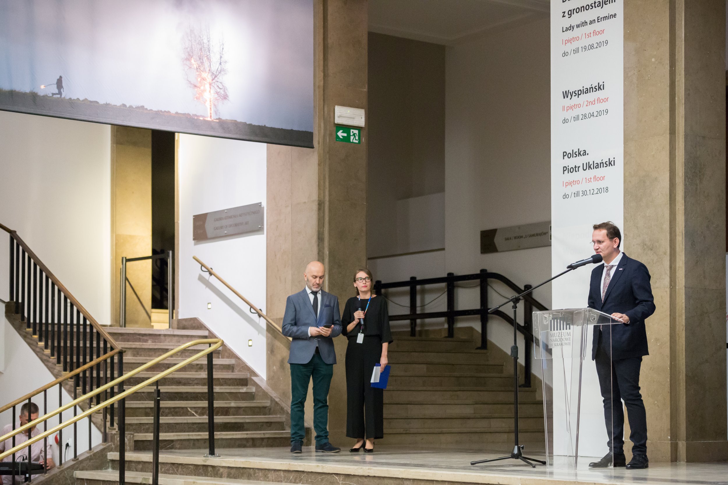 Wnętrze muzeum z szerokimi marmurowymi schodami i dużym banerem fotograficznym nad wejściem, na podwyższeniu stoją trzej mówcy — mężczyzna przy przezroczystym pulpicie przemawiający do mikrofonu, obok mężczyzna w marynarce patrzący na telefon oraz kobieta w czarnym stroju trzymająca mikrofon i teczkę, a po prawej słup z pionową tablicą informacyjną o wystawach.