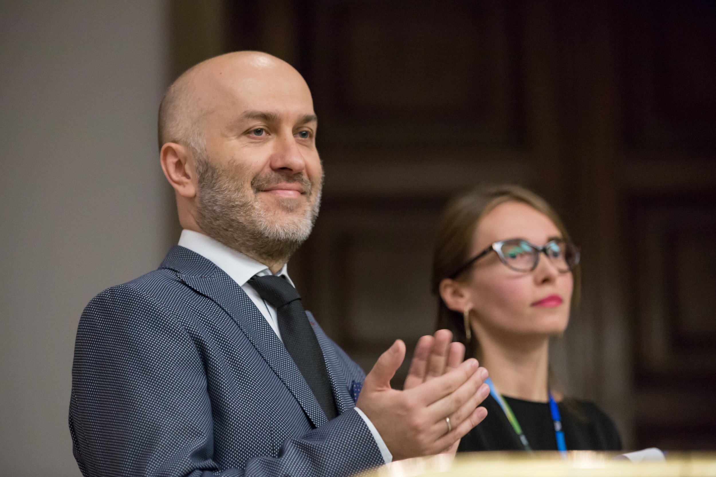 Na pierwszym planie stojący mężczyzna z łysą głową i siwą brodą w granatowym garniturze w drobny wzór, białej koszuli i czarnym krawacie klaska dłońmi i ma delikatny uśmiech; w tle po prawej lekko rozmyta kobieta w okularach, ciemnym ubraniu i identyfikatorze na smyczy.