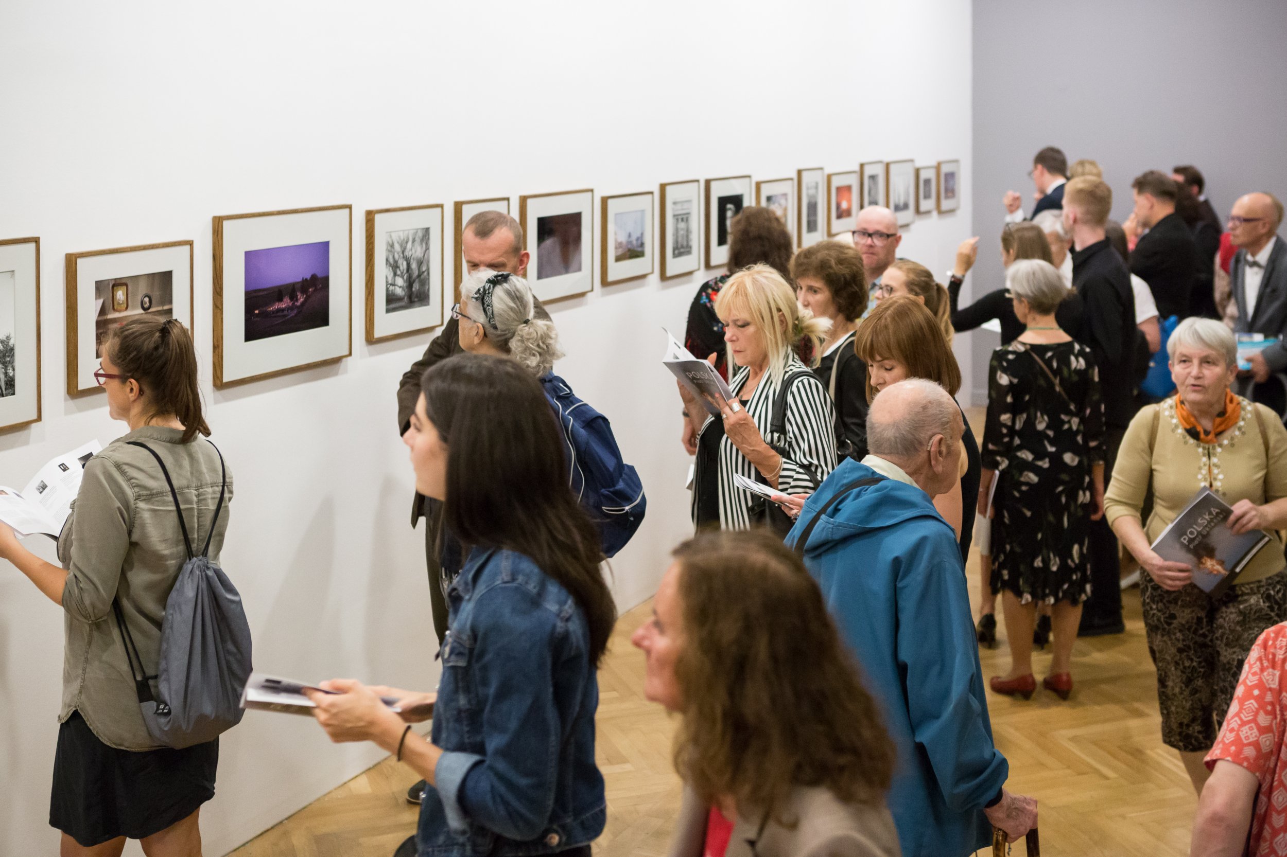 Wnętrze galerii z długą białą ścianą, na której w równym rzędzie wiszą małe oprawione fotografie, oraz grupą zwiedzających w różnym wieku stojących blisko ściany, przeglądających katalogi i oglądających prace na jasnym drewnianym parkiecie.