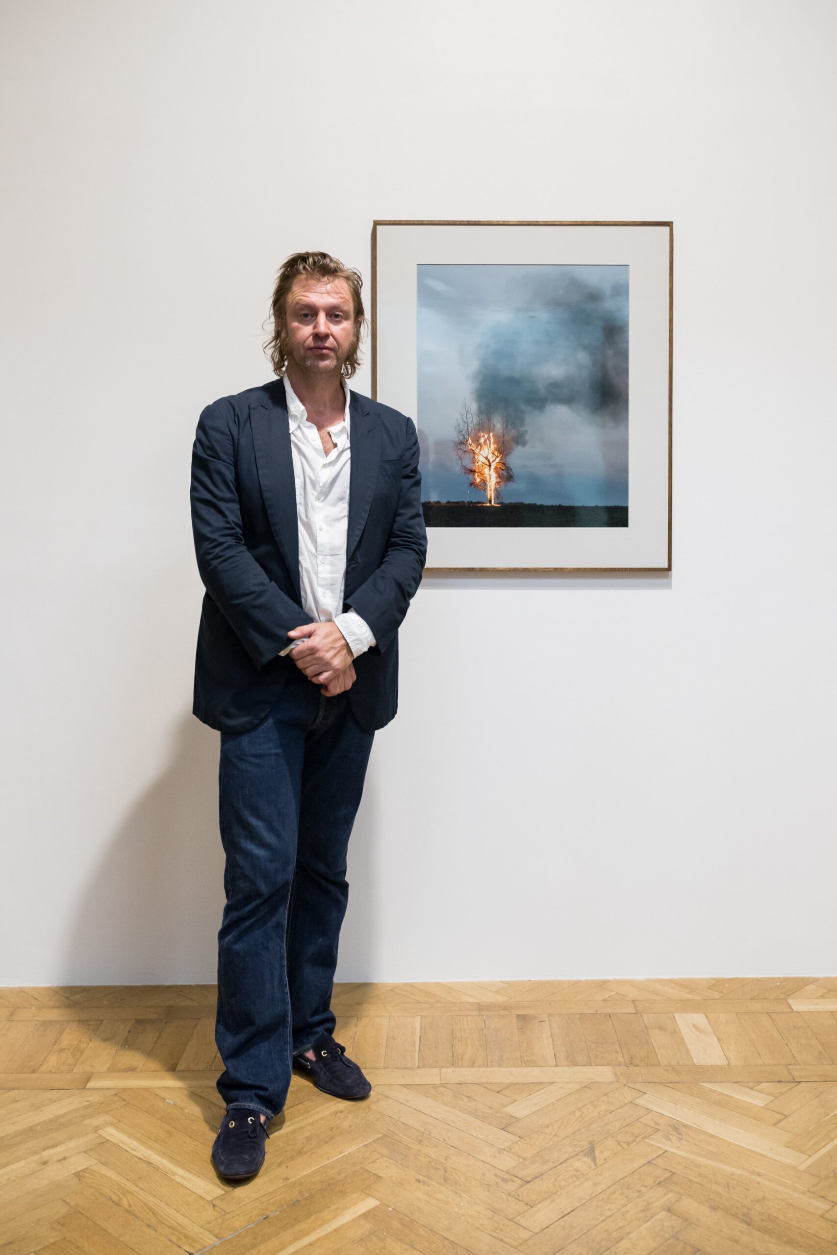 Mężczyzna stojący na drewnianej podłodze przed białą ścianą w galerii, ubrany w ciemną marynarkę, białą koszulę i dżinsy, z rękami złożonymi przed sobą, obok oprawionego zdjęcia przedstawiającego samotne drzewo płonące i unoszący się dym na tle niebiesko‑szarego nieba.
