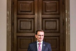 Mężczyzna w ciemnym garniturze, białej koszuli i różowym krawacie, w okularach i z przypinką na klapie, stoi na tle dużych ciemnobrązowych drewnianych drzwi z dekoracyjnymi panelami.