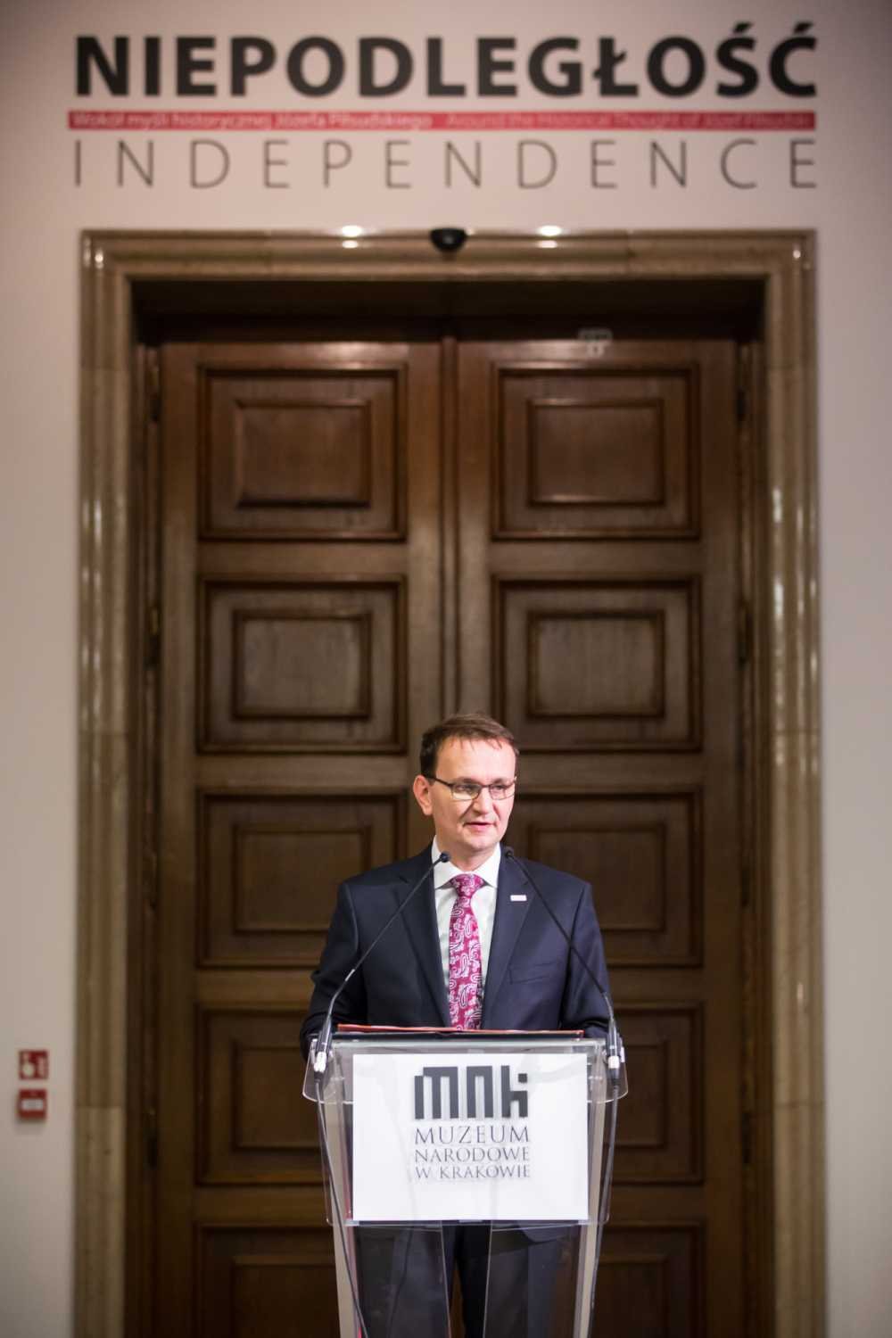 Mężczyzna w ciemnym garniturze i wzorzystym krawacie stoi za przezroczystą mównicą z mikrofonami przed dużymi drewnianymi drzwiami pod napisem NIEPODLEGŁOŚĆ INDEPENDENCE, a na froncie mównicy widoczne logo Muzeum Narodowe w Krakowie.