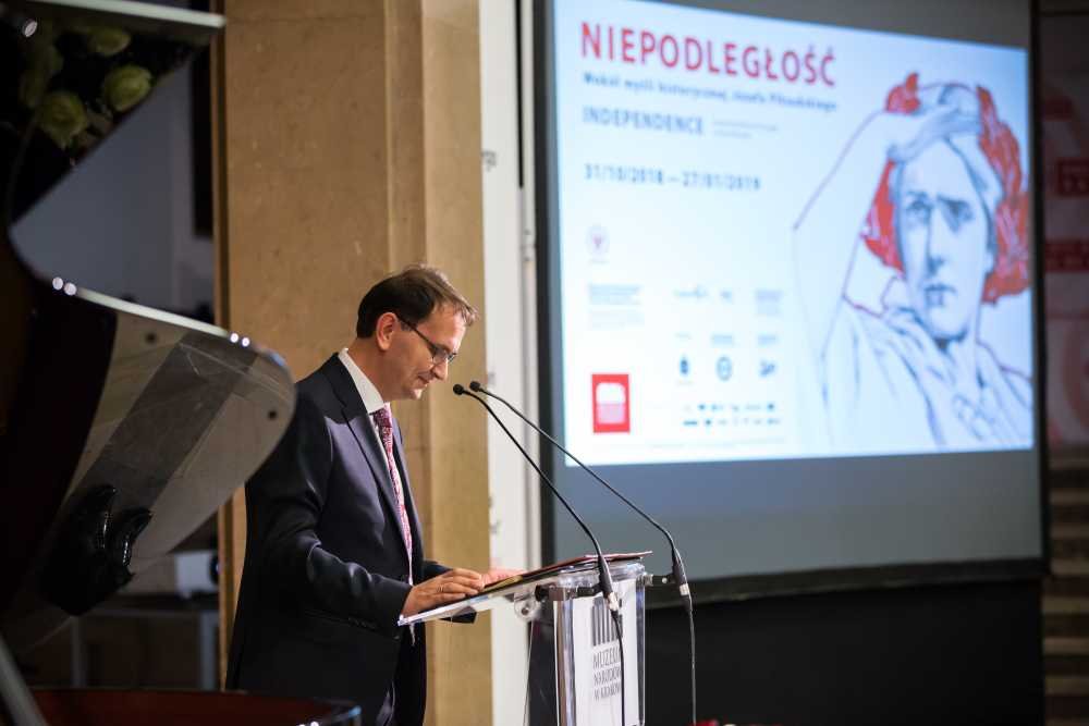 Mężczyzna w okularach i garniturze stoi przy przezroczystym pulpicie z mikrofonami czytając z dokumentów, po lewej widać fragment fortepianu, a w tle duży ekran z napisem "NIEPODLEGŁOŚĆ" i rysunkiem głowy postaci klasycznej.