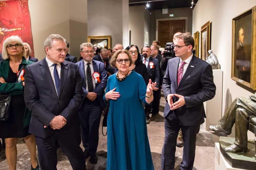 Grupa elegancko ubranych osób przechodząca przez salę muzealną z obrazami i rzeźbami; na pierwszym planie kobieta w turkusowej sukni i okularach gestykuluje, obok niej po lewej i prawej stoją mężczyźni w garniturach, w tle inni uczestnicy (niektórzy z biało‑czerwonymi kotylionami) oraz osoba z kamerą.