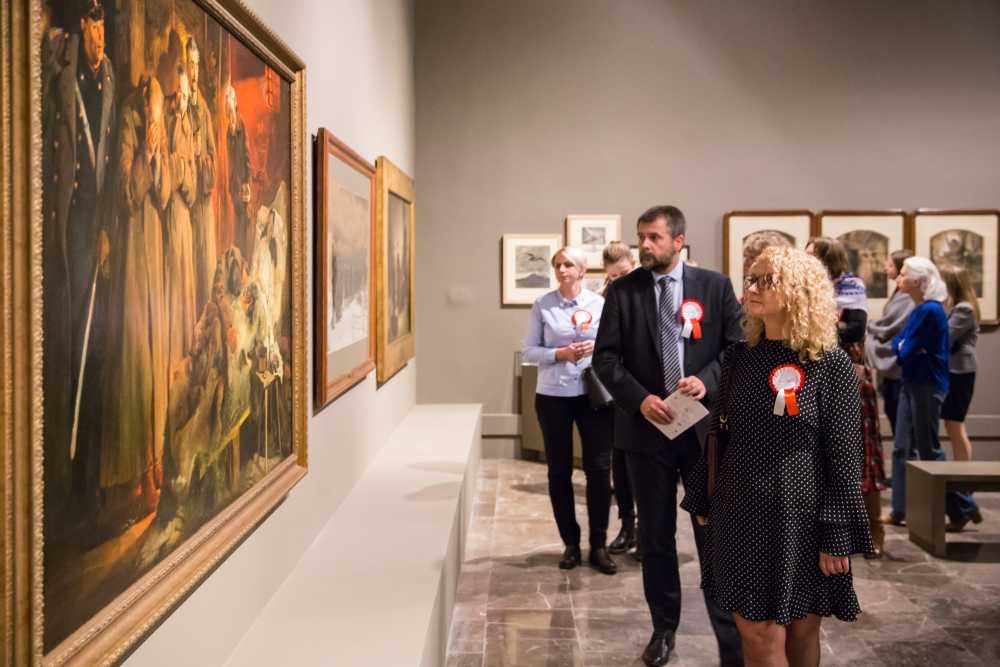 Wnętrze galerii z dużym oprawionym obrazem po lewej i mniejszymi pracami na ścianie, po prawej grupa zwiedzających — w tym mężczyzna w garniturze i kobieta z kręconymi blond włosami w czarnej sukience w białe kropki, niektórzy z nich mają przypięte czerwono-białe rozetki.