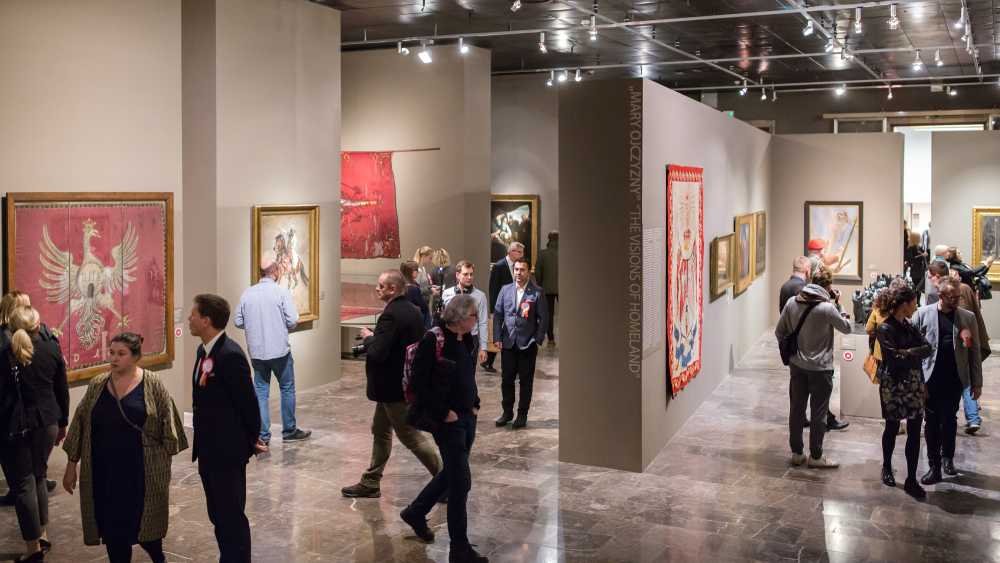 Wnętrze galerii sztuki z działowymi jasnoszarymi ściankami, na których wiszą obrazy i gobeliny, środkowa ścianka z pionowym napisem "THE VISIONS OF HOMELAND", oraz liczne osoby spacerujące i oglądające eksponaty na błyszczącej kamiennej podłodze pod sufitowym oświetleniem.