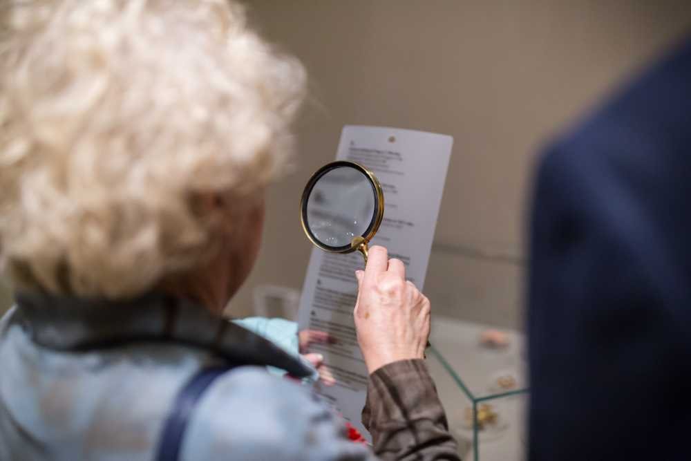 Osoba z siwymi, kręconymi włosami widziana od tyłu trzyma lupę o złotej oprawie nad kartką z drobnym tekstem przy szklanej gablocie, po prawej widać fragment innej osoby w ciemnym ubraniu.
