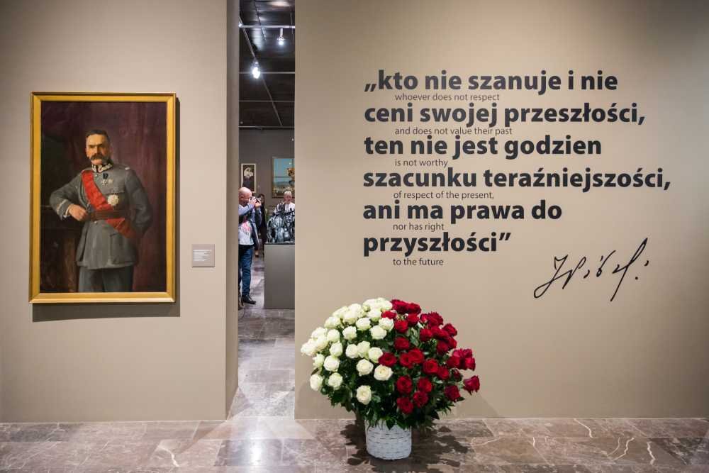Widok sali wystawowej z lewej oprawionym portretem mężczyzny w historycznym mundurze, pośrodku prześwit do sąsiedniej sali z widocznymi zwiedzającymi, po prawej dużym napisem w języku polskim z mniejszymi angielskimi tłumaczeniami i podpisem, oraz na pierwszym planie koszem czerwonych i białych róż ustawionym na marmurowej podłodze.