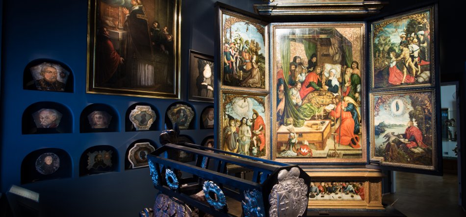 Wnętrze muzeum z otwartym, bogato malowanym tryptykiem przedstawiającym liczne sceny z postaciami religijnymi; na pierwszym planie ozdobna drewniano-metalowa balustrada z herbem, w tle nisze ścienne z portretowymi medalionami i dużym obrazem w ramie.