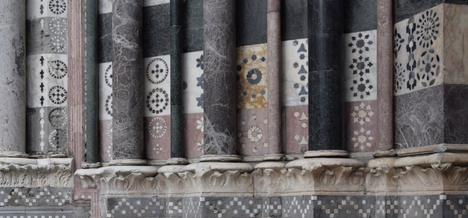 Fragment dekoracyjnej elewacji kamiennej z rzędem pionowych cylindrycznych kolumn z marmuru w odcieniach czerni, szarości i różu, między którymi umieszczone są prostokątne panele z okrągłymi rozetami mozaikowymi, nad rzeźbionym gzymsem i pasem geometrycznych płytek.