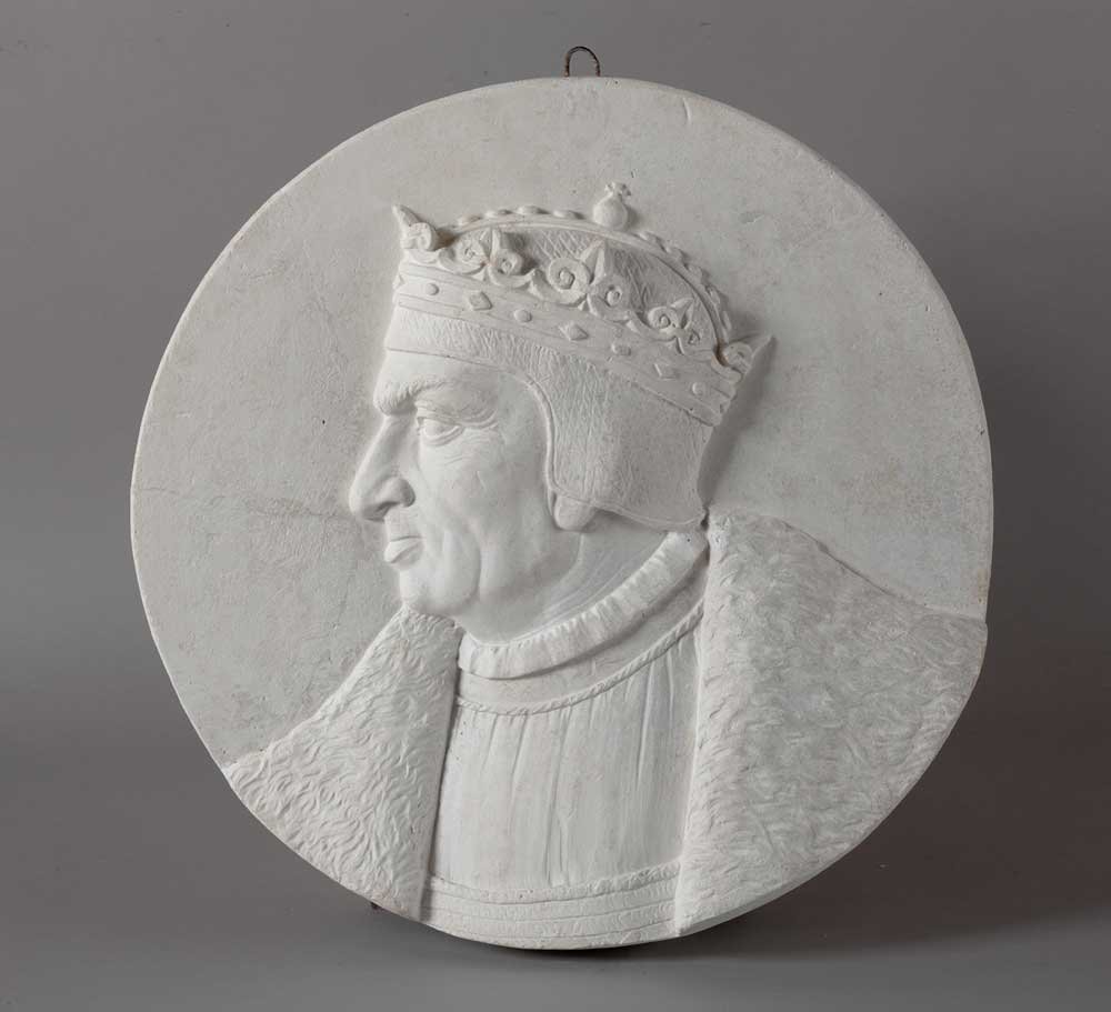 Okrągły biały relief przedstawiający profil mężczyzny zwróconego w lewo w zdobionej koronie i futrzanym płaszczu, ustawiony na gładkim szarym tle z małym metalowym zawieszkiem u góry.