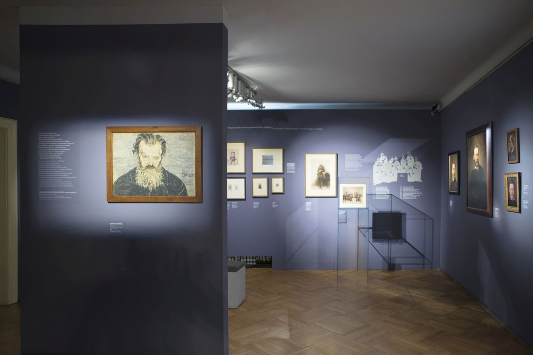 Wnętrze galerii z granatowymi ścianami i drewnianą podłogą; po lewej na przegrodzie oświetlony rysunek brodatego mężczyzny w drewnianej ramie, w głębi po prawej kilka oprawionych fotografii i portretów, przeszklona gablotka ekspozycyjna oraz tablice z opisami na ścianach.
