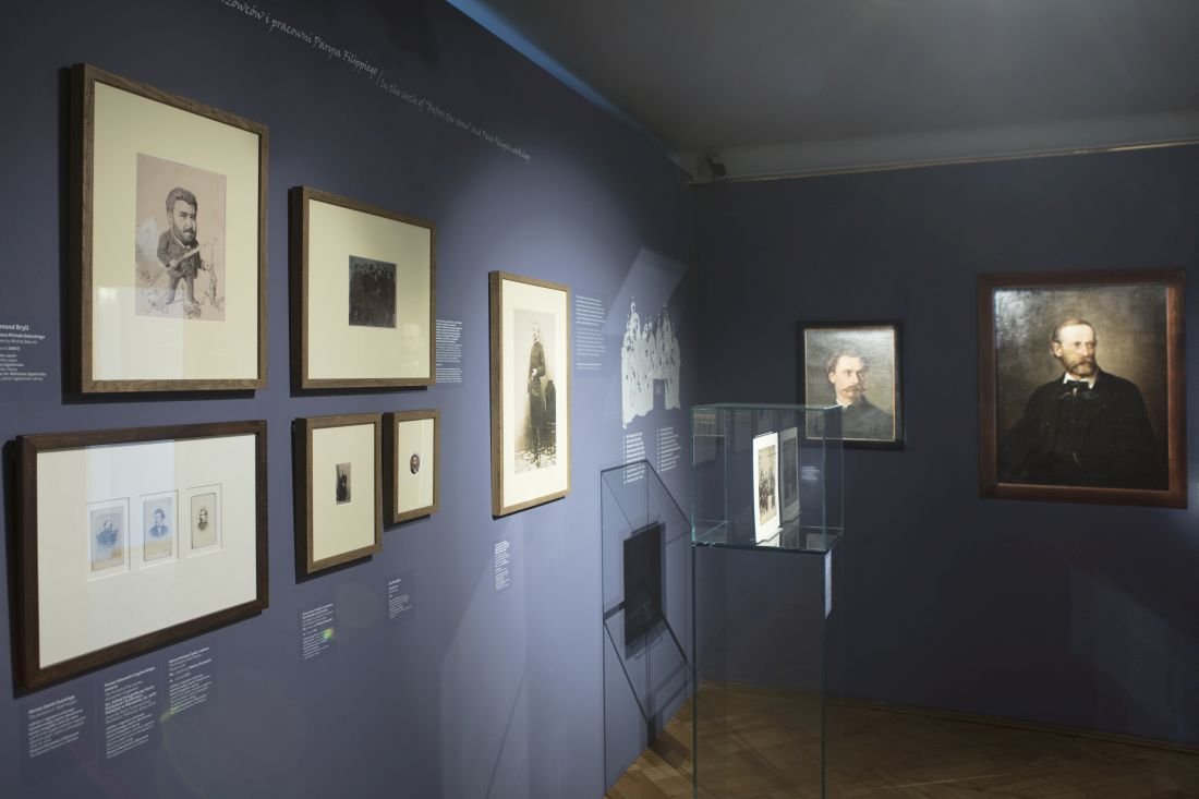Wnętrze galerii z ciemnoniebieską ścianą, na której po lewej w grupie wiszą oprawione fotografie i szkice, na środku duży pionowy zdjęcie, na środku pomieszczenia przezroczysta przeszklona gablotka z eksponatem, a po prawej dwie większe oprawione prace, wszystko oświetlone punktowo i opatrzone krótkimi opisami tekstowymi przy ramach.